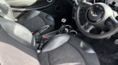 Interior of a MINI Cooper showing the front seats, steering wheel, and dashboard. The seats are black leather, and the driver's seat has a manual gear shift in front. The dashboard features a prominent circular speedometer, air vents, and a steering wheel with the MINI logo.