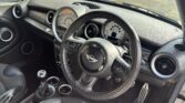The image shows the interior of a MINI Cooper car, focusing on the driver's side. The dashboard features a large, circular speedometer and various controls. The steering wheel has the MINI logo in the center. The seats are black leather, and there is a manual gear shift with three cup holders nearby.