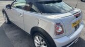 A silver MINI compact car with a black roof and rear spoiler is parked on a gray asphalt surface. The car has a yellow license plate with the registration number "YF63 XXE." In the background, several other vehicles and parts of buildings can be seen.