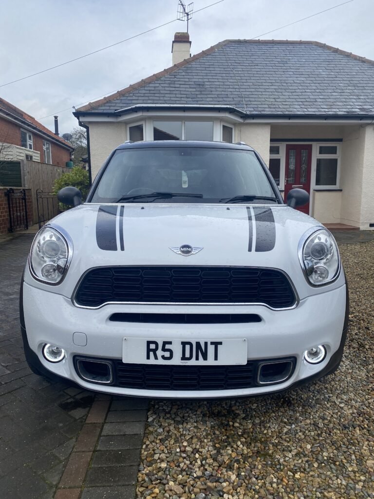 A white MINI Cooper with black racing stripes on the hood is parked on a gravel driveway in front of a single-story house. The car's license plate reads "R5 DNT." The house has a grey roof, a small front yard, and a red front door.