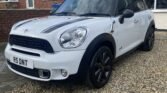 A white MINI Cooper with a black roof and black wheels is parked on a gravel driveway in front of a brick house with a well-kept garden. The license plate reads "R5 DNT." The sky is partly cloudy, providing soft, diffused light over the scene.