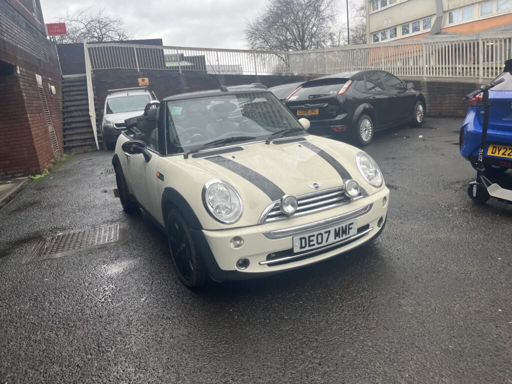 A cream MINI Cooper convertible with black racing stripes is parked on wet pavement near a brick building. The license plate reads "DE07 MMF." Two other vehicles—a black car and a blue car with a yellow plate—are nearby. Trees and a fence are visible in the background.