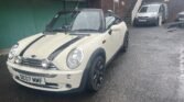 A white MINI Cooper convertible with black racing stripes is parked on a wet, asphalt surface outside a building with brown brick walls and green garage doors. The car, registration number DE07 MMF, has its black soft top down. In the background, a white van is also parked.