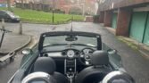 A MINI convertible with its top down is parked on a wet, dark asphalt road. The car's interior features two seats, a detailed dashboard with gauges, and a gear shift. Nearby, an electric scooter stands by. In the background, you'll find brick buildings, green lawns, trees, and a staircase.