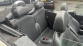 Interior of a MINI convertible with dark leather seats and metallic roll bars behind the headrests. The backseat is visible, featuring stitching details and seat belt buckles. Raindrops are visible on the glass. In the background, other parked vehicles are partially visible near a brick wall.