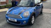 A blue MINI Cooper with black racing stripes is parked on a residential street in front of red-brick houses. The car has a license plate reading "YS11 HKO." There is a moving truck in the background. The sky is partly cloudy with sunlight illuminating the scene.