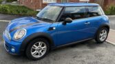 A blue MINI Cooper with black trim is parked on a residential street, facing left, with slightly tinted windows. Behind the car stands a red brick, two-story home featuring white trim and brown roofs. The paved street has a small patch of greenery on the left side.