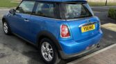 A blue MINI Cooper is parked on a paved area near a large building. The car has a yellow rear license plate reading "Y511 HKO." The sky is partly cloudy, and a few shrubs are visible near the building.