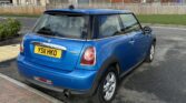 A MINI Cooper D, with a striking blue finish, is parked on a road beside a brick pavement. The yellow registration plate reads "YS11 HKO." Industrial-style buildings with corrugated metal walls and an overcast sky loom in the background, while patches of greenery spot the area near the buildings.