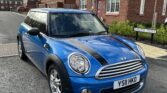 A blue MINI Cooper with black racing stripes is parked on the side of a residential street. The car's license plate reads "YS11 HKO". The background features two-story brick houses with pitched roofs, white-trimmed windows, and a cloudy sky overhead. A signpost reads "Calorim Drive".