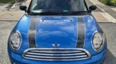 A blue MINI Cooper is parked on a residential street, featuring black racing stripes on the hood and a European "YS11 HKO" license plate. The street is lined with modern brick houses and small trees. The sky is partly cloudy, and sunlight casts shadows on the car.