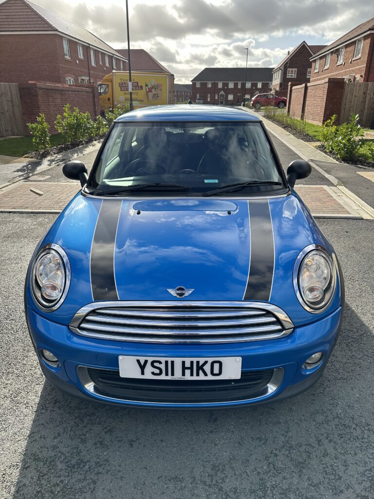 A blue MINI Cooper is parked on a residential street, featuring black racing stripes on the hood and a European "YS11 HKO" license plate. The street is lined with modern brick houses and small trees. The sky is partly cloudy, and sunlight casts shadows on the car.
