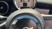 A view from the driver's seat of a car, showcasing a black steering wheel adorned with the MINI logo in the center. The speedometer behind it reads zero. Part of the dashboard and a ventilation outlet are visible, with a brick building and its windows serving as the backdrop.