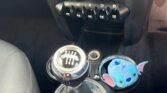 A car interior focusing on the gear shift area of a MINI. The gear shift is manual, with a stitched black boot. To the right, there is a blue stuffed toy resembling Disney's Stitch. The dashboard includes an audio system and a row of toggle switches beneath it. The car seats and floor are black.