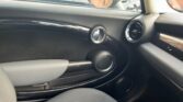 The image shows the interior of a MINI, focusing on the passenger side. It features a sleek, black door with unique circular design elements. The air vent and part of the dashboard are visible, along with a portion of the gray passenger seat. The window and side mirror are also partially visible.