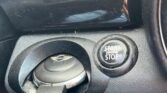 Close-up of a MINI ignition area featuring a key partially inserted into the ignition slot positioned below a circular push-button labeled "START ENGINE STOP". The dashboard is black with some reflective elements, and a gloved hand appears to be holding the key.