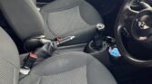 Interior of a small black mini car, showing two front seats with grey and black upholstery. The dashboard, steering wheel, gear stick, and center console are visible. Outside the window, part of another blue car is visible. The floor has minimal clutter.