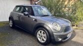 A gray MINI Cooper with a sleek design is parked on a partly mossy driveway in front of a white garage door. The car features silver alloy wheels and a sunroof. Bushes and a brick house with a brown tiled roof enhance the background. License plate visible.