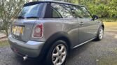 A gray MINI One is parked on a wet, paved driveway. The car features a black roof and silver alloy wheels, positioned at an angle showcasing its rear and right side. The backdrop includes trees and greenery, hinting at a suburban setting.