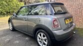 A grey MINI Cooper is parked in a driveway beside a brick wall. The car features a sloping rear design, silver alloy wheels, and a rear license plate that reads "PL 04 WGU." Surrounded by greenery and trees, the setting provides a natural backdrop. The MINI's windows are tinted.