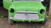 A green vintage MINI with no headlights or bumper is parked in a garage. The front grille is exposed, and the left door is open, revealing part of the interior. The word "HOONIGAN" is visible on the upper windshield. The ground is uneven with patches of grass and gravel.
