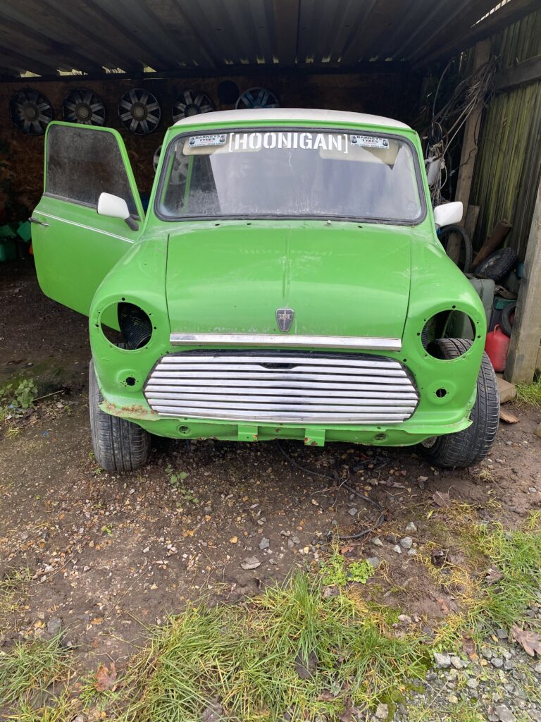 A green vintage MINI with no headlights or bumper is parked in a garage. The front grille is exposed, and the left door is open, revealing part of the interior. The word "HOONIGAN" is visible on the upper windshield. The ground is uneven with patches of grass and gravel.