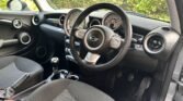 Interior of a car showing the dashboard, steering wheel with the MINI logo, and control panel with large circular displays. The front passenger and driver seats are upholstered in grey fabric. The gear stick and handbrake are visible between the seats. Outside greenery is visible through windows.