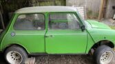 A classic green MINI with a white roof is parked in a wooden shed. The car shows significant wear, with missing wheels and a partially rusted body. Various automotive parts and debris are scattered inside and around the shed. A British Royal Air Force roundel is on the rear side panel.
