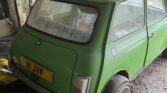 An old, green vintage MINI car with a slightly dusty exterior is parked inside a shed with a partially open door. The car has a license plate reading "A1 JVP" and features circular side mirrors and a classic British roundel symbol on the side. Various tools and items are scattered around the shed.