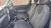 This image shows the interior of a small MINI car with black and grey fabric seats. The front seats are bucket-style, and the dashboard features a large central speedometer, an infotainment system, and several control knobs. The black steering wheel complements the cupholders in the center console.