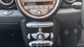 Close-up view of a MINI car's dashboard and central console. The dashboard features a large speedometer in the center, measuring speed in MPH. Below are various controls, including climate control dials, audio system buttons, and toggle switches. The gearshift knob with a six-speed manual pattern is in the foreground.