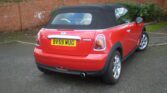 A red MINI convertible Cooper S with a black soft top is parked on a paved surface near a brick wall and greenery. The vehicle's license plate reads "BV59 WUG." The rear view showcases its taillights, exhaust pipe, and silver alloy wheels.