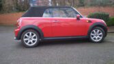A red MINI Cooper convertible is parked on a residential street. The car has a black soft-top roof and silver alloy wheels. The background features a red-brick wall, bare trees, and a distant blue fence. The car is oriented in side profile, showcasing the MINI's compact design.