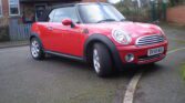 A red MINI Cooper convertible parked near a brick building with a tiled roof. The car has a black soft top, silver alloy wheels, and a UK license plate reading "BV59 WUG." The background shows a wooden fence, shrubbery, and a partially visible tree. The ground is paved with some wear.