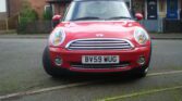 A red MINI Cooper with a British license plate "BV59 WUG" is parked on a residential street. The car is angled slightly to the left, with houses and a garden fence visible in the background. The front grille and headlights are prominent, and the pavement around the car is clear.