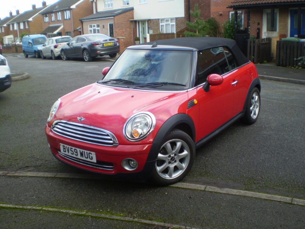 A red MINI Cooper convertible with its black soft top up is parked on a residential street. The vehicle has shiny silver alloy wheels and a UK registration plate reading "BV59 WUG." Brick houses with cars in driveways are visible in the background.