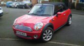 A red MINI Cooper convertible with its black soft top up is parked on a residential street. The vehicle has shiny silver alloy wheels and a UK registration plate reading "BV59 WUG." Brick houses with cars in driveways are visible in the background.