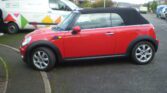 A red convertible MINI Cooper is parked on the side of a residential street. The car has a black soft top roof. In the background, a white van with various colorful geometric shapes on its side is parked on the street. The street is lined with houses and greenery.