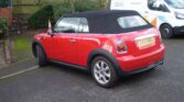 A red MINI Cooper convertible with a black soft top is parked on a street. The rear license plate reads "BV53 VJG." To the right, a white van with a "Maintenance" sign is visible, and in the background, there is a wooden fence and some greenery. The road surface is asphalt.