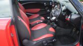 The image shows the interior of a red MINI with black and red seats. The driver's seat and passenger seat have red side bolsters and black central sections. The dashboard is black with chrome-rimmed dials and a central speedometer. The steering wheel is black. The door is open, revealing an outside brick wall.
