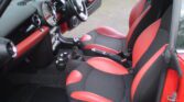 The image shows the interior of a MINI with two-tone seats in black and red. The dashboard is stylish, featuring a large, circular speedometer at its center. The car has a manual transmission, and the door panels match the red and black color scheme. The floor mats appear to be clean and well-maintained.