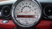 Close-up view of a MINI car speedometer and central control panel. The speedometer displays a range up to 160 mph, with a digital clock in the center reading 2:51. Below the speedometer are buttons labeled "Audio" and "Menu" on a stylish red and black dashboard.