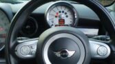 A close-up of a MINI Cooper's steering wheel and dashboard. The steering wheel boasts a prominent MINI logo at its center, with control buttons on both spokes. Behind it, a circular speedometer reads 12°C and shows a range of 252 miles. The background reveals greenery through the car's windshield.
