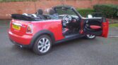 A red convertible MINI Cooper is parked on a paved surface with the driver's door open. The interior, including the steering wheel and seats, is visible. The car features a black fabric roof, which is folded down, and a yellow license plate reads "DV59 WUG." A red brick wall and bushes are in the background.