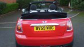 A red MINI Cooper convertible with its black top down is parked on a paved area. The vehicle's rear license plate reads "BV59 WUG." The car is surrounded by greenery, including bushes and a grassy area, with a brick wall and wooden fence in the background.
