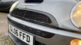 Close-up of a grey MINI Cooper with a focus on the front grille and license plate, which reads "GL06 FFD." The car's headlights are on, and it is parked next to another vehicle with a brick building and pavement visible in the background. The MINI emblem is prominently displayed above the grille.