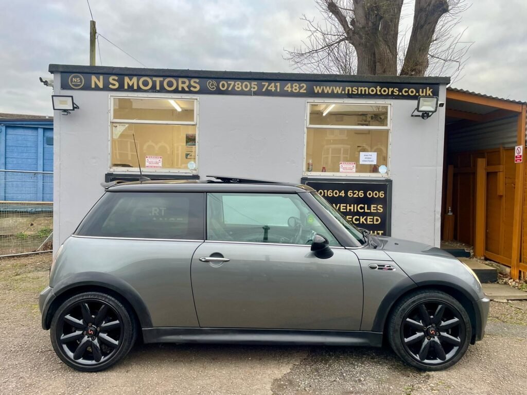 A silver MINI Cooper with black rims is parked in front of a small car dealership building. The building has a grey facade and displays the name "NS Motors" with contact information in bold yellow letters on a black sign above the windows and door.