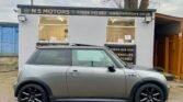 A silver MINI Cooper with black rims is parked in front of a small car dealership building. The building has a grey facade and displays the name "NS Motors" with contact information in bold yellow letters on a black sign above the windows and door.