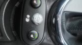 Close-up of a MINI's center console showcasing seat heater controls. The console features two round buttons with seat icons and green indicator lights, plus a central control dial for adjusting mirror positions. The right side highlights part of a chrome-trimmed black leather gear shift.