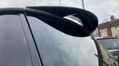 Close-up view of a black MINI's rear spoiler under an overcast sky. The spoiler is mounted on the vehicle's trunk, slightly raised, and features a sleek, aerodynamic design. In the background, there are blurry houses, power lines, and another car.